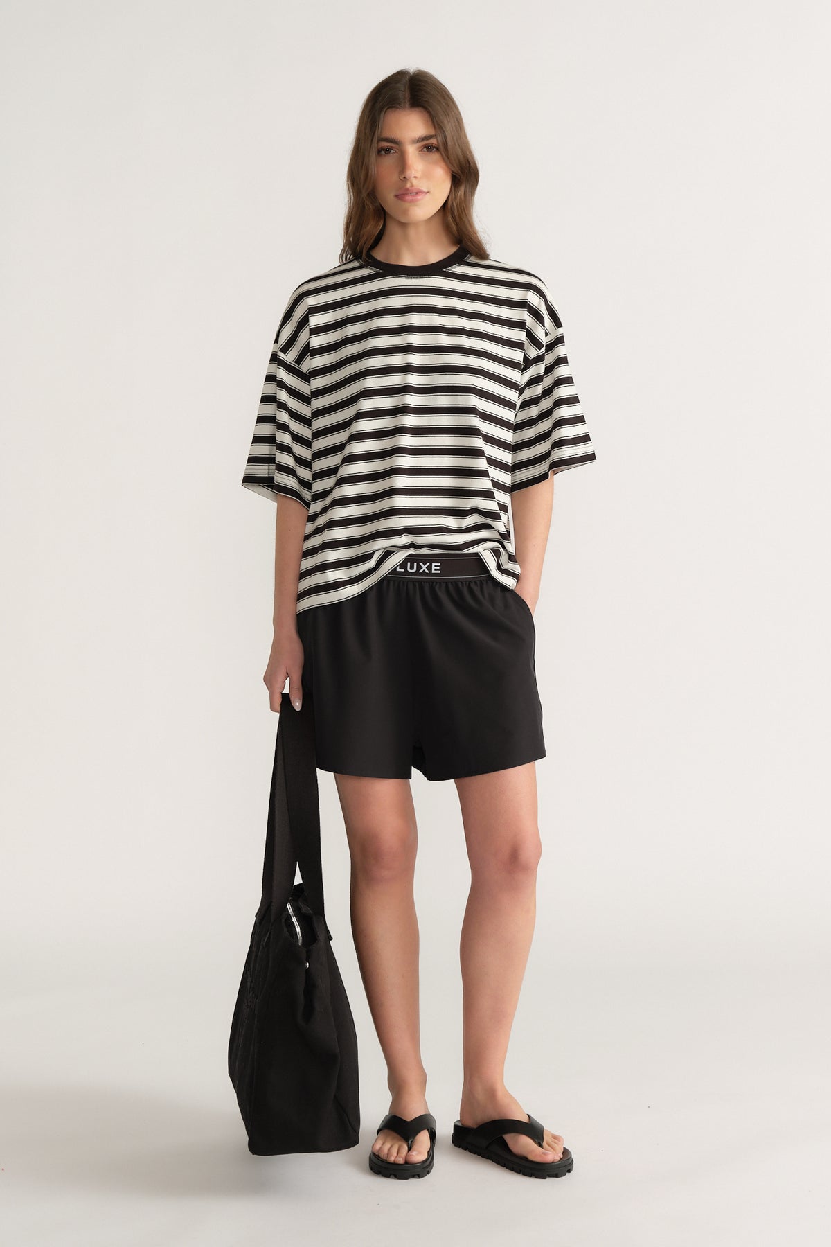 Woman wearing an oversized black and white striped tee with black shorts, sandals and a tote bag