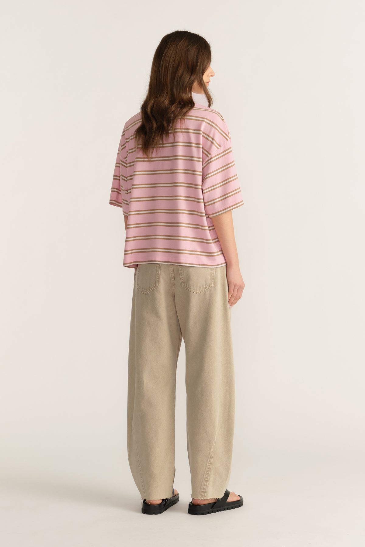 Woman wearing an oversized pink striped tee with beige pants and black sandals, viewed from behind