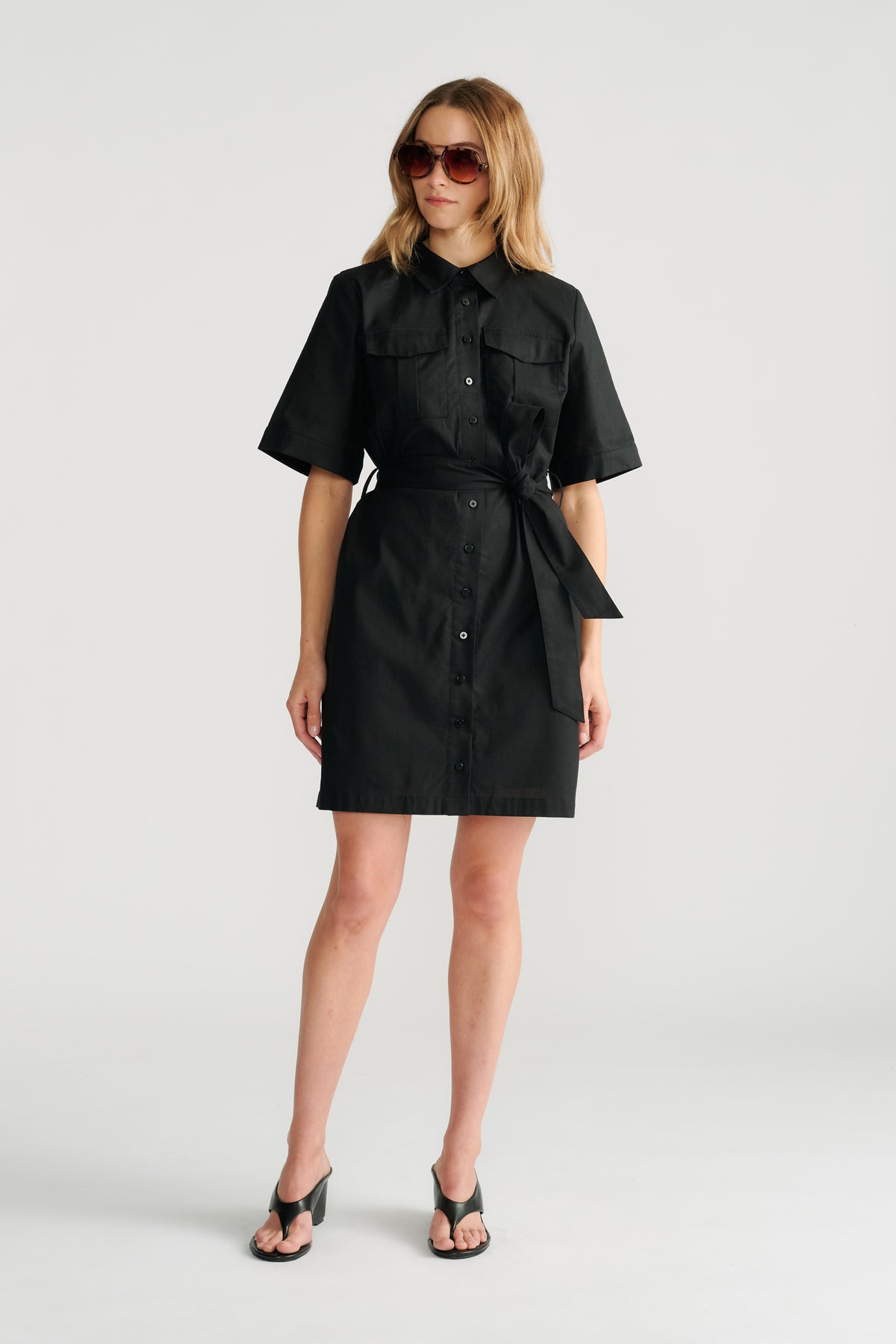 Woman wearing a black belted short sleeve mini dress with front buttons and oversized sunglasses styled with black heels.