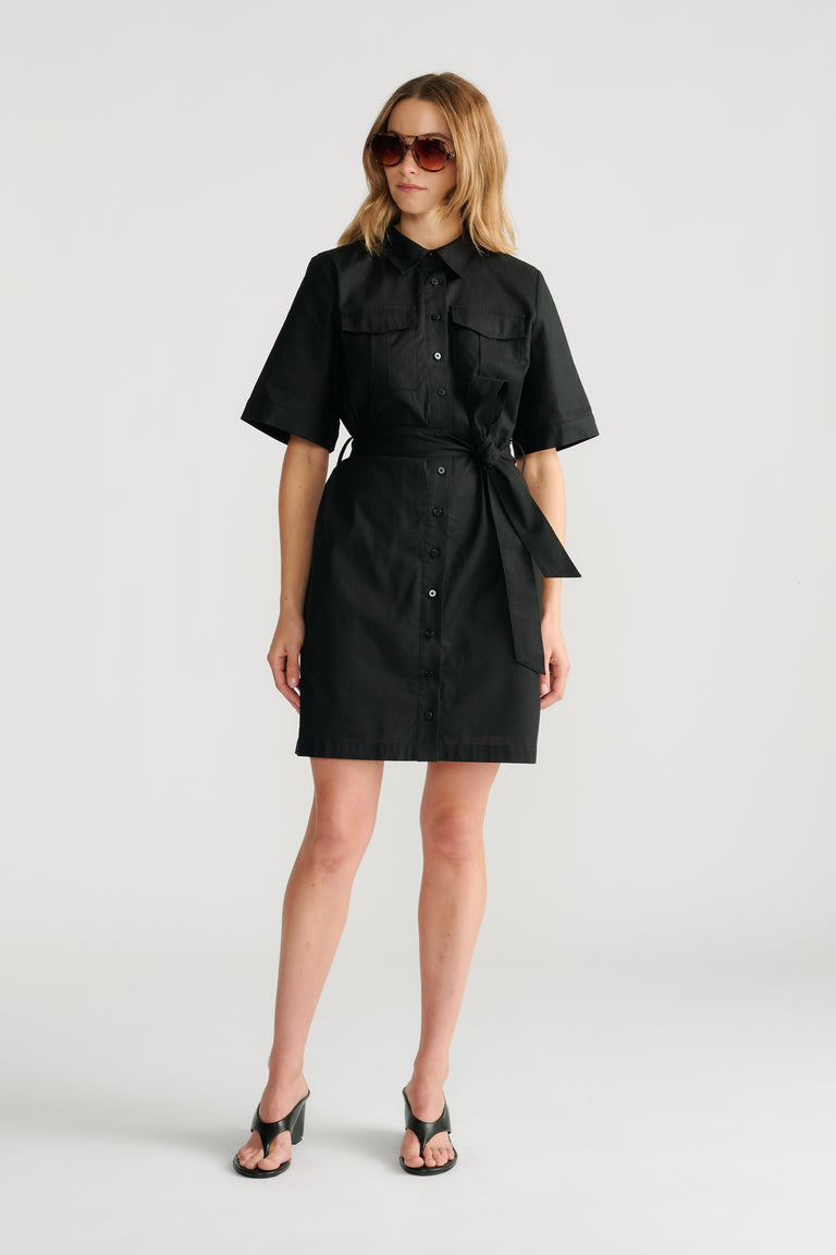 Woman wearing a black belted short sleeve mini dress with front buttons and oversized sunglasses styled with black heels.