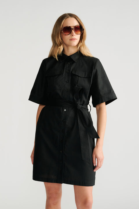 Woman wearing a black short sleeve belted dress with button details and oversized sunglasses, standing against a plain background.