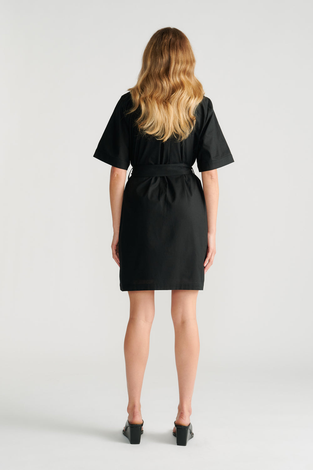 Woman wearing a black short sleeve mini dress with a belted waist, shown from the back with loose wavy hair.