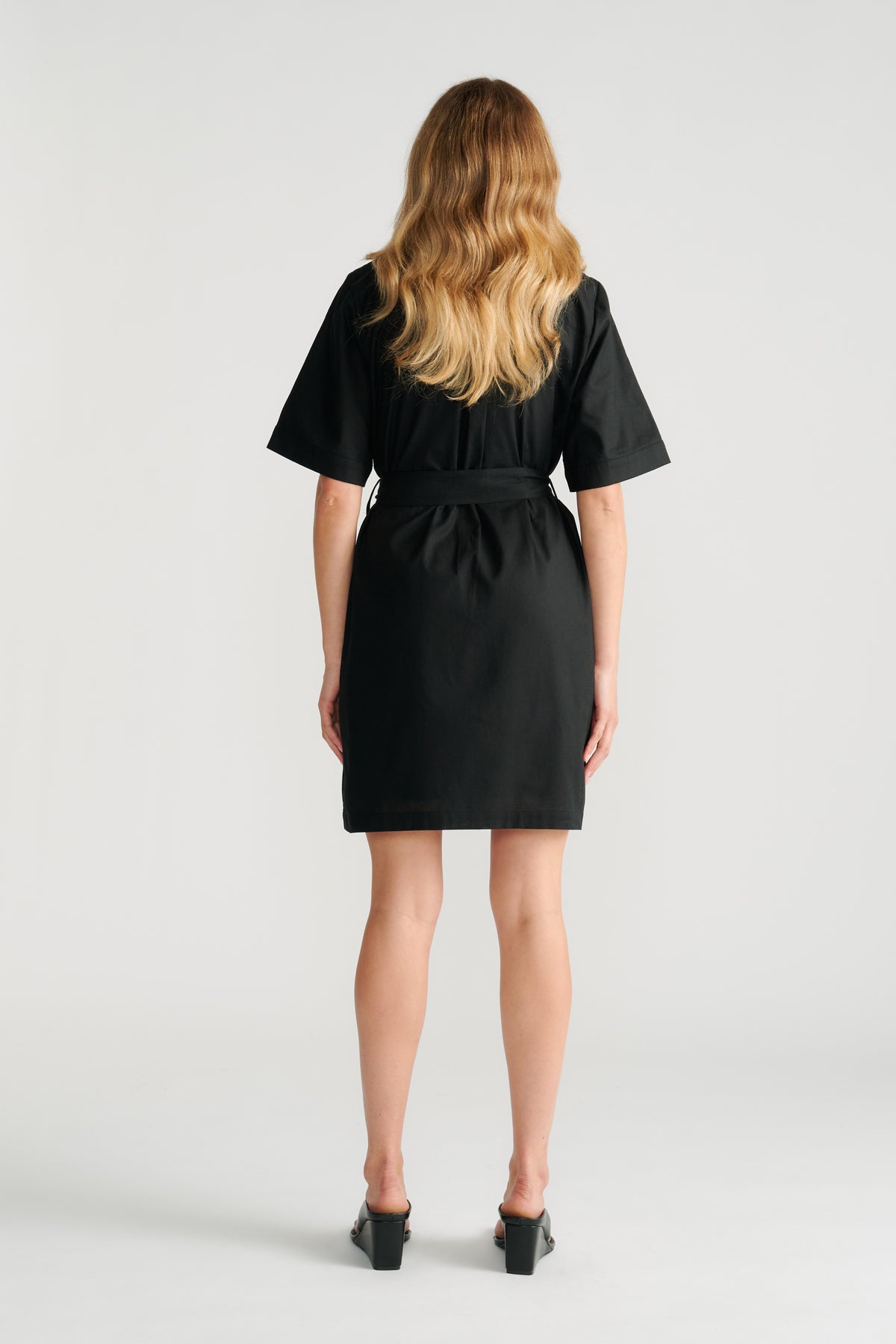 Woman wearing a black short sleeve mini dress with a belted waist, shown from the back with loose wavy hair.