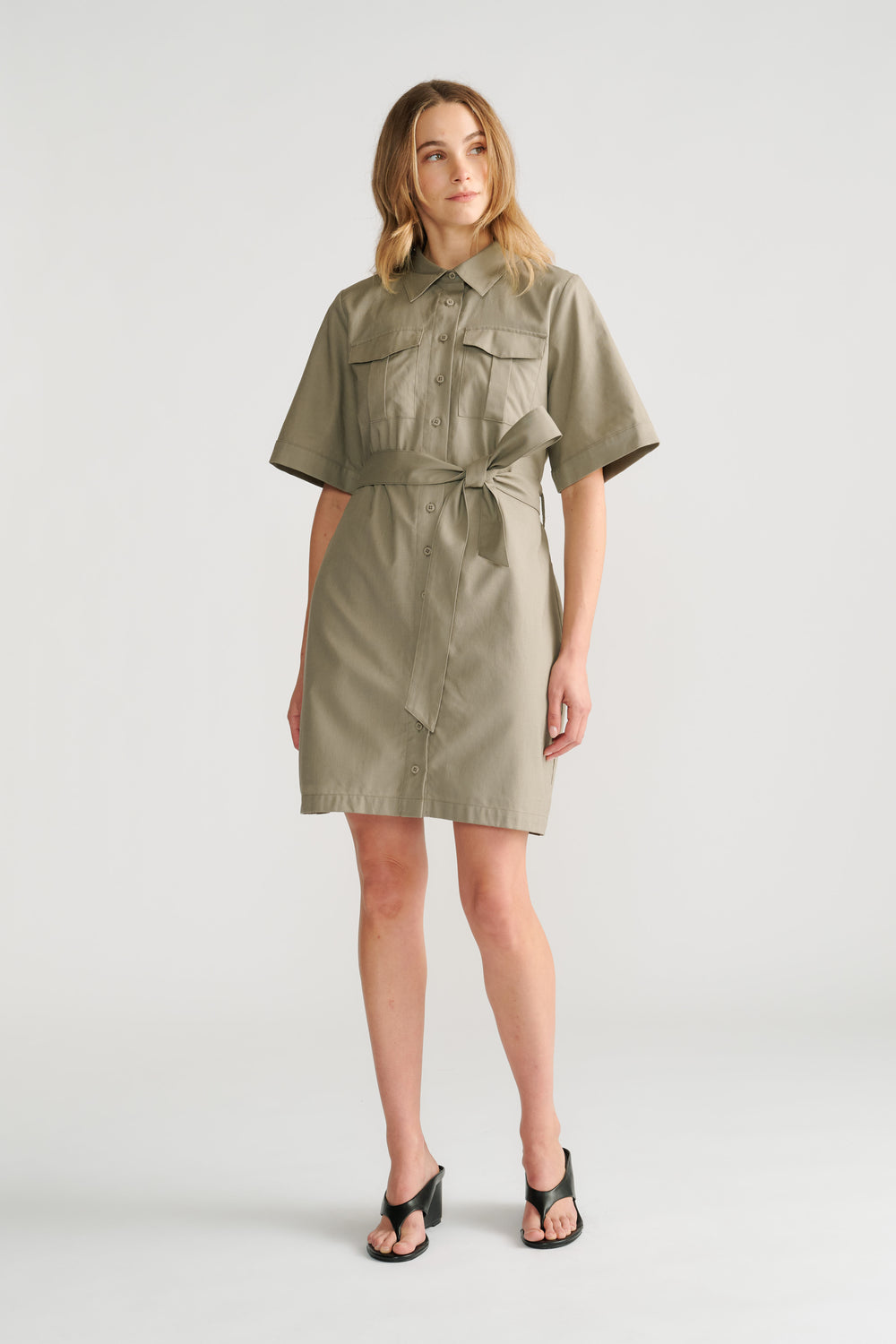 Woman wearing a sage green belted mini dress with short sleeves and button-front detail paired with black heeled sandals.