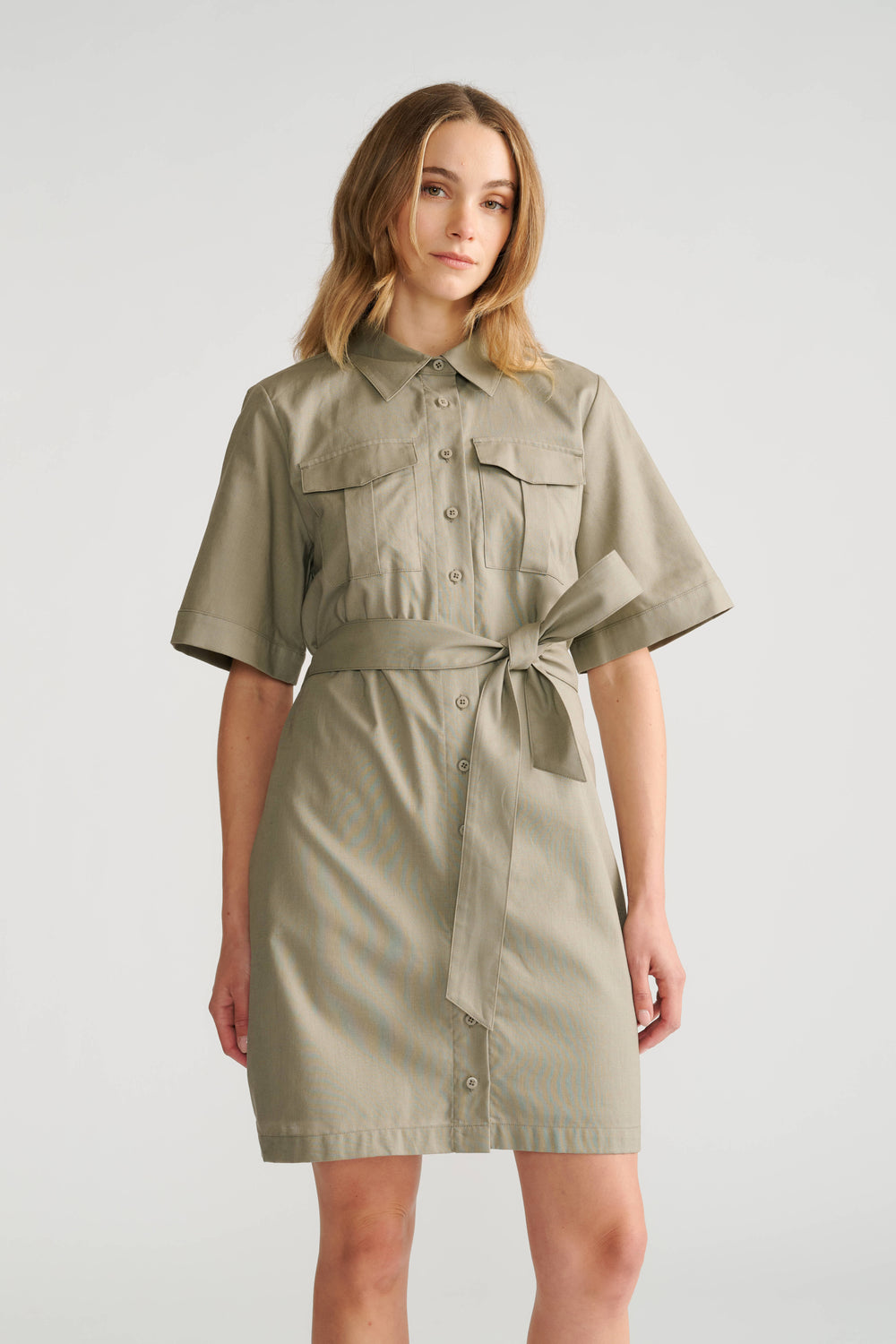 Woman wearing a sage green short sleeve mini dress with chest pockets and a waist tie detail.
