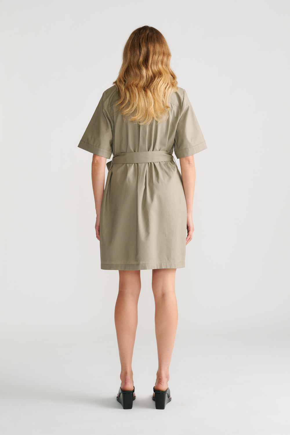 Woman seen from behind wearing a sage green belted short sleeve mini dress with black block heel shoes.