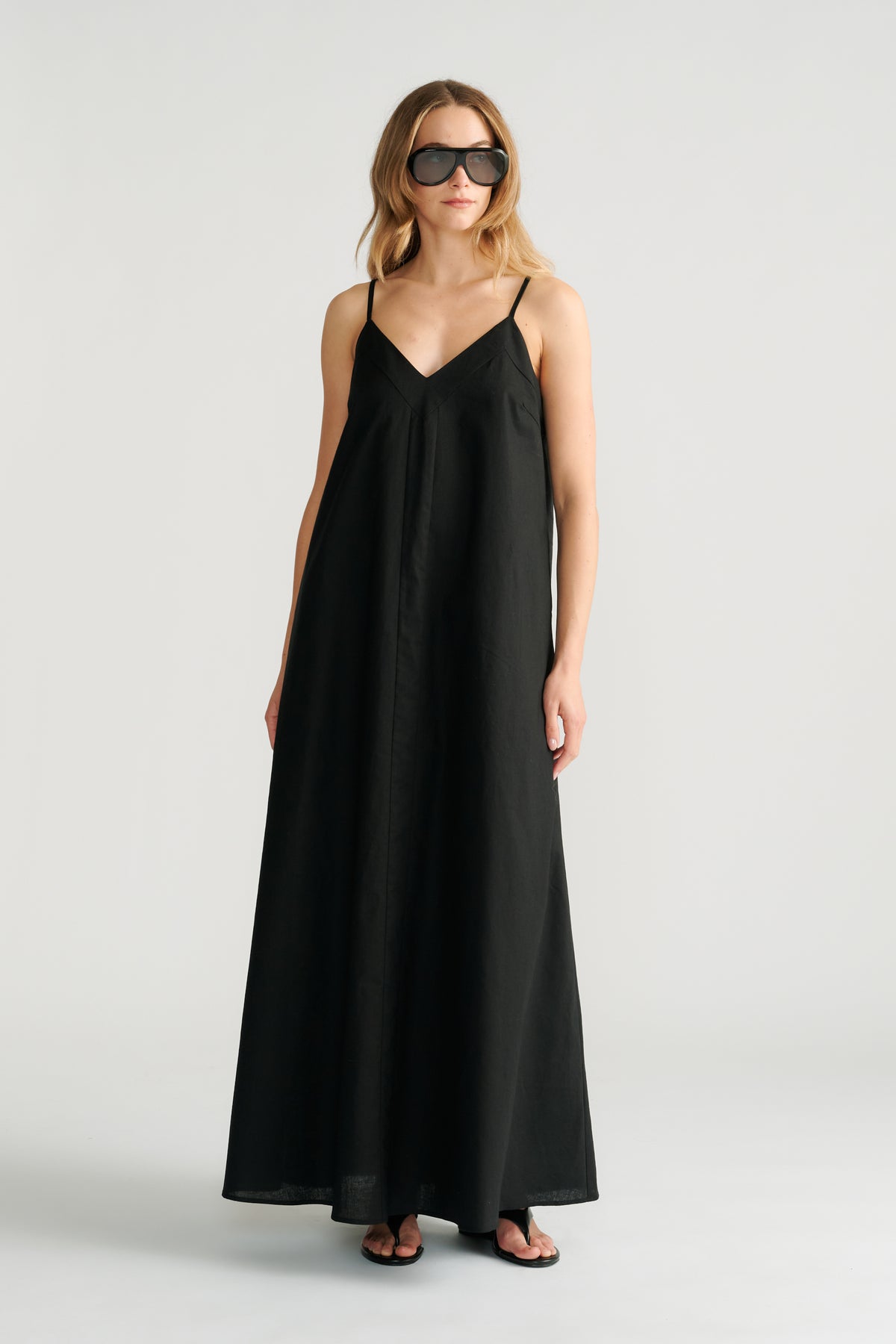 Woman wearing a flowing black maxi dress with thin straps and a V-neckline paired with oversized sunglasses and sandals.