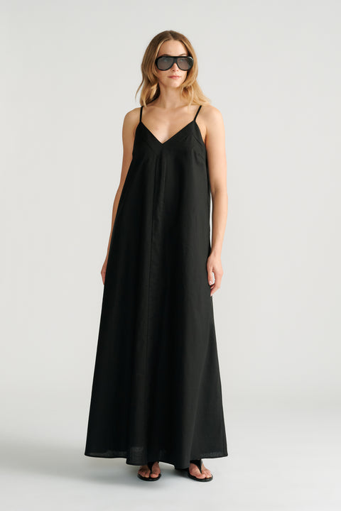 Woman wearing a flowing black maxi dress with thin straps and a V-neckline paired with oversized sunglasses and sandals.