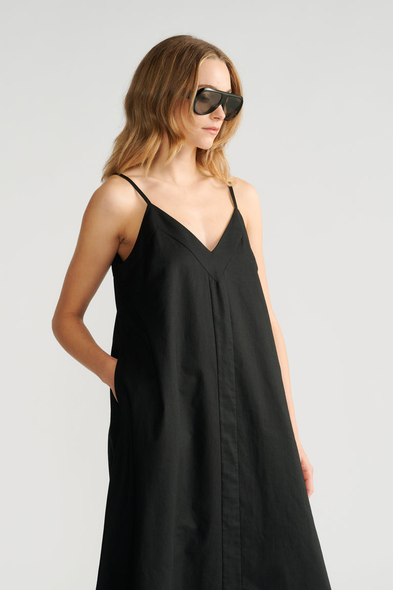 Woman wearing a black sleeveless trapeze dress with thin straps and side pockets, paired with oversized sunglasses.