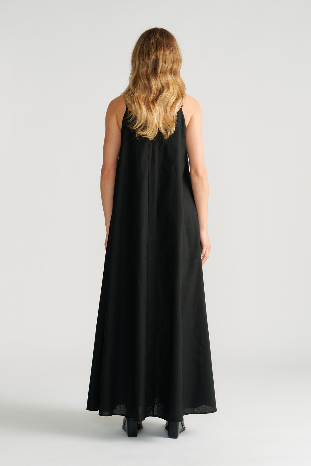 Woman wearing a flowing black maxi dress with thin straps, shown from the back with wavy hair and black heeled shoes.
