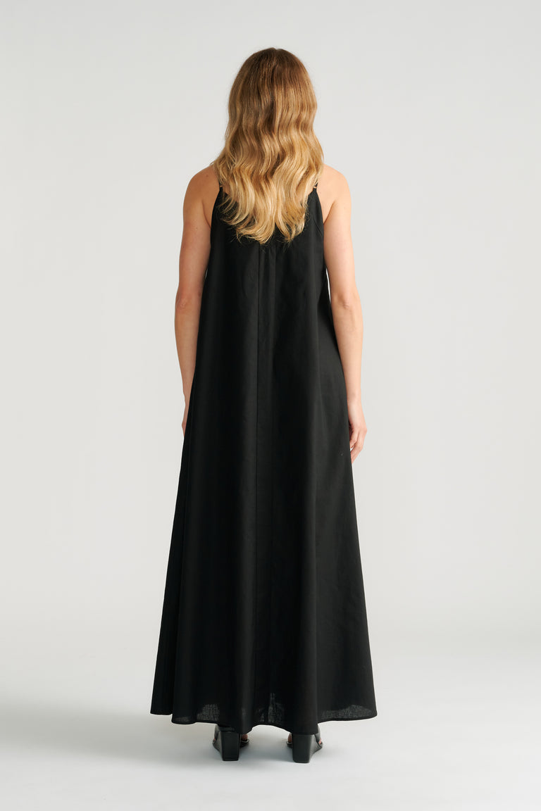 Woman wearing a flowing black maxi dress with thin straps, shown from the back with wavy hair and black heeled shoes.