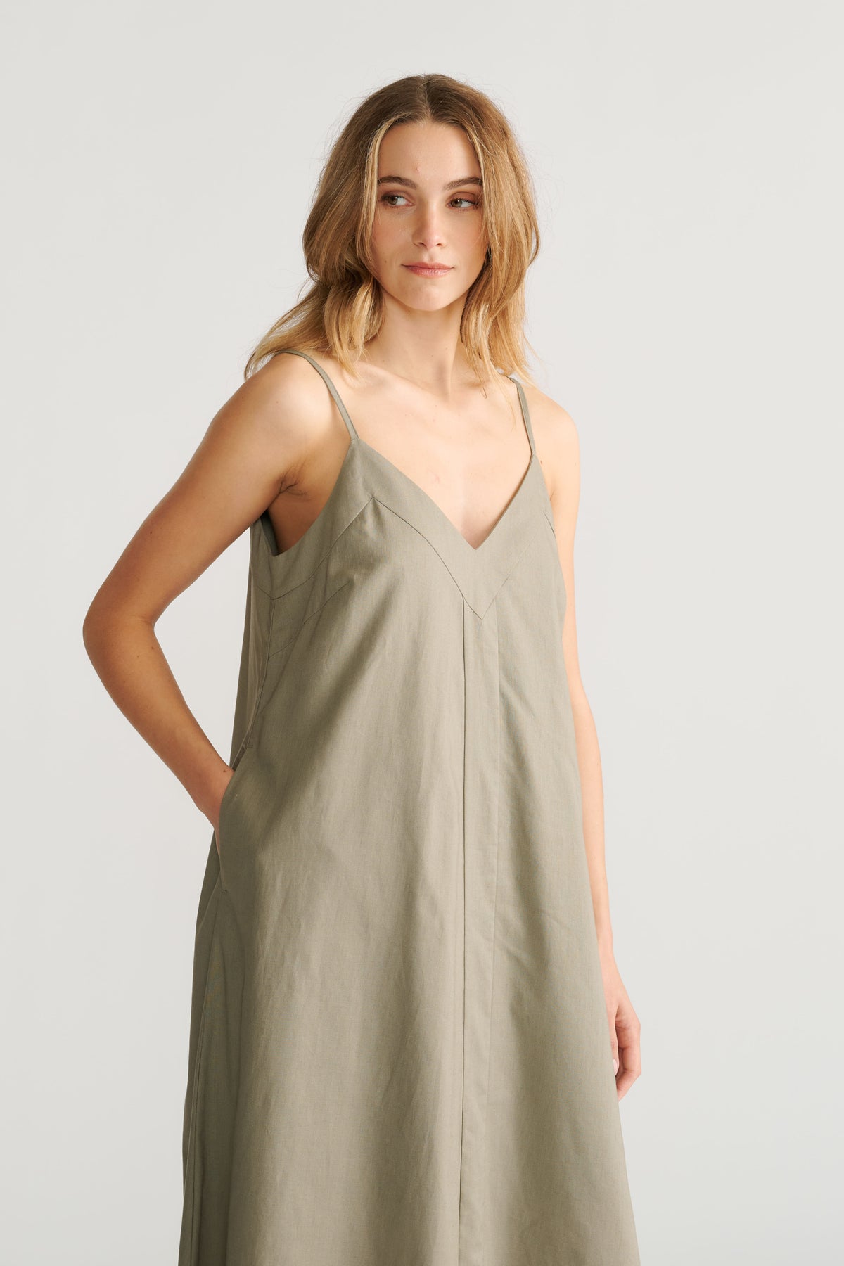 Woman wearing a sage green trapeze maxi dress with thin straps and a V-neckline, posing with a relaxed expression.