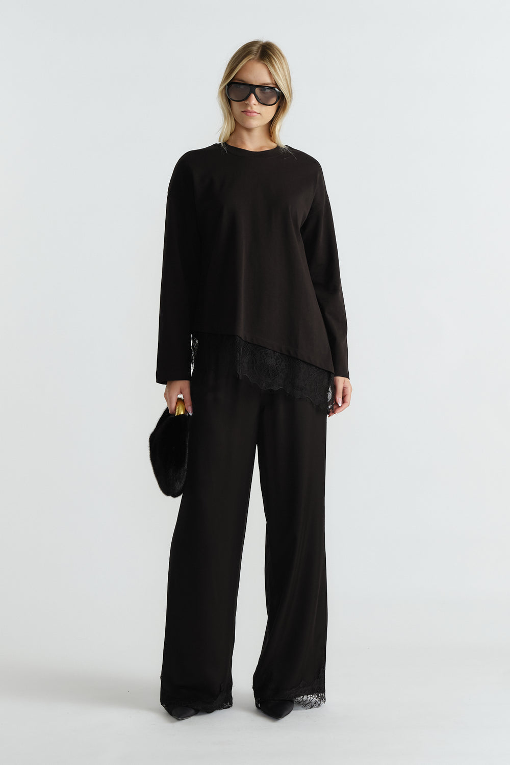 Woman wearing a black long sleeve top with lace trim and matching wide-leg pants, carrying a black fur clutch.