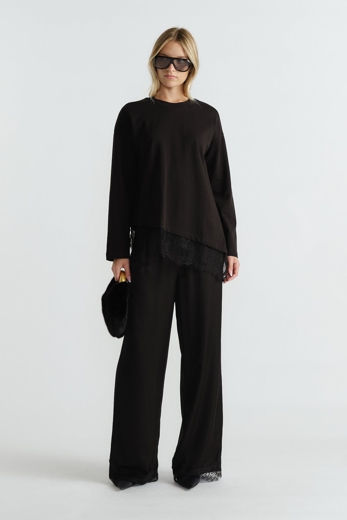 Woman wearing a black long sleeve top with lace trim and matching wide-leg pants, carrying a black fur clutch.