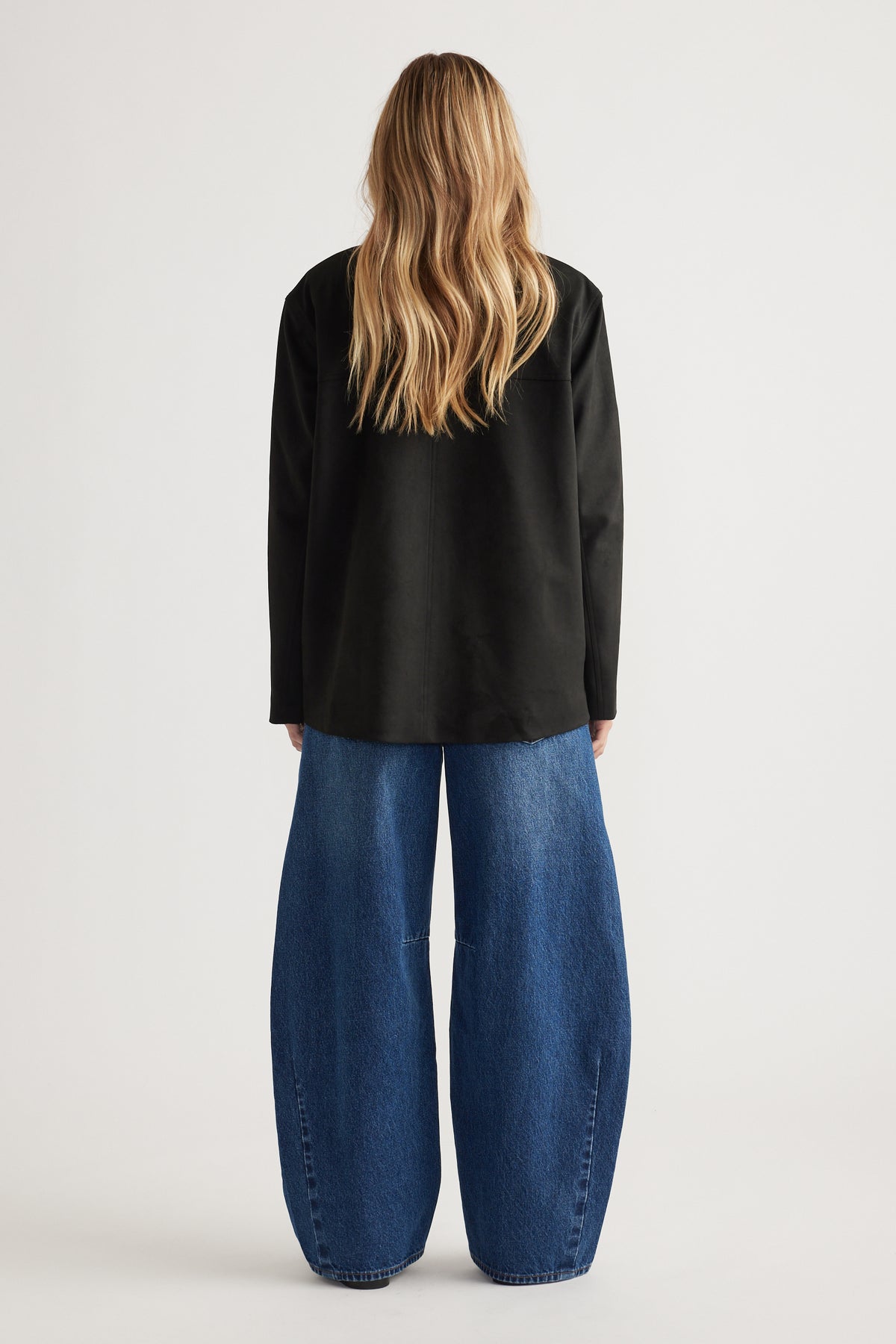 Woman wearing a relaxed black coat paired with wide-leg blue denim jeans, shown from the back.