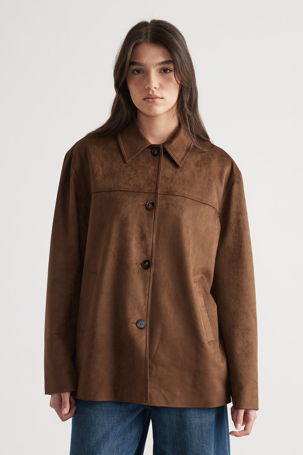 Woman wearing a rich brown button-up coat with a classic collar and relaxed fit over casual blue jeans.