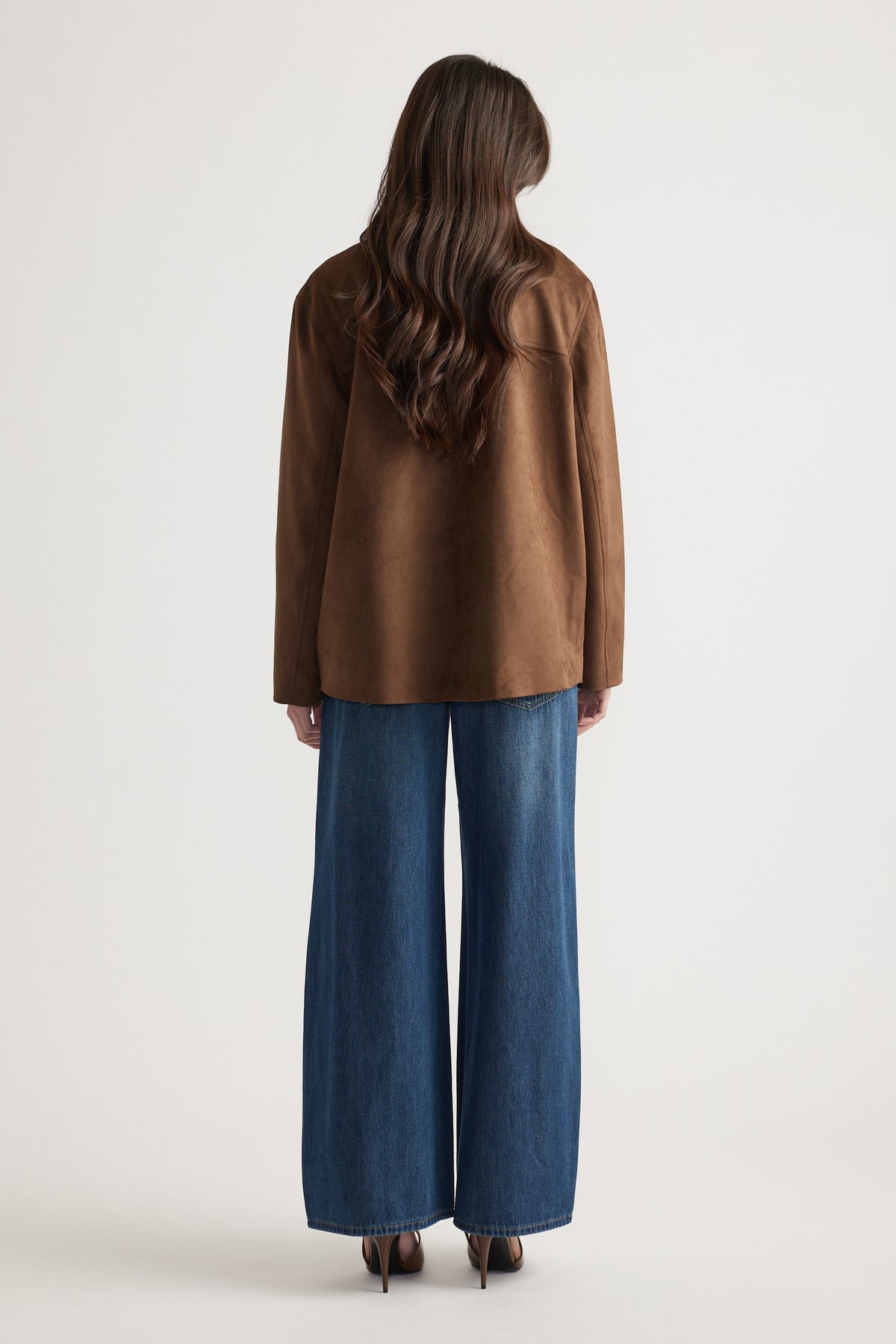 Woman with long wavy hair wearing a bronze car coat and wide-leg denim jeans, shown from behind.