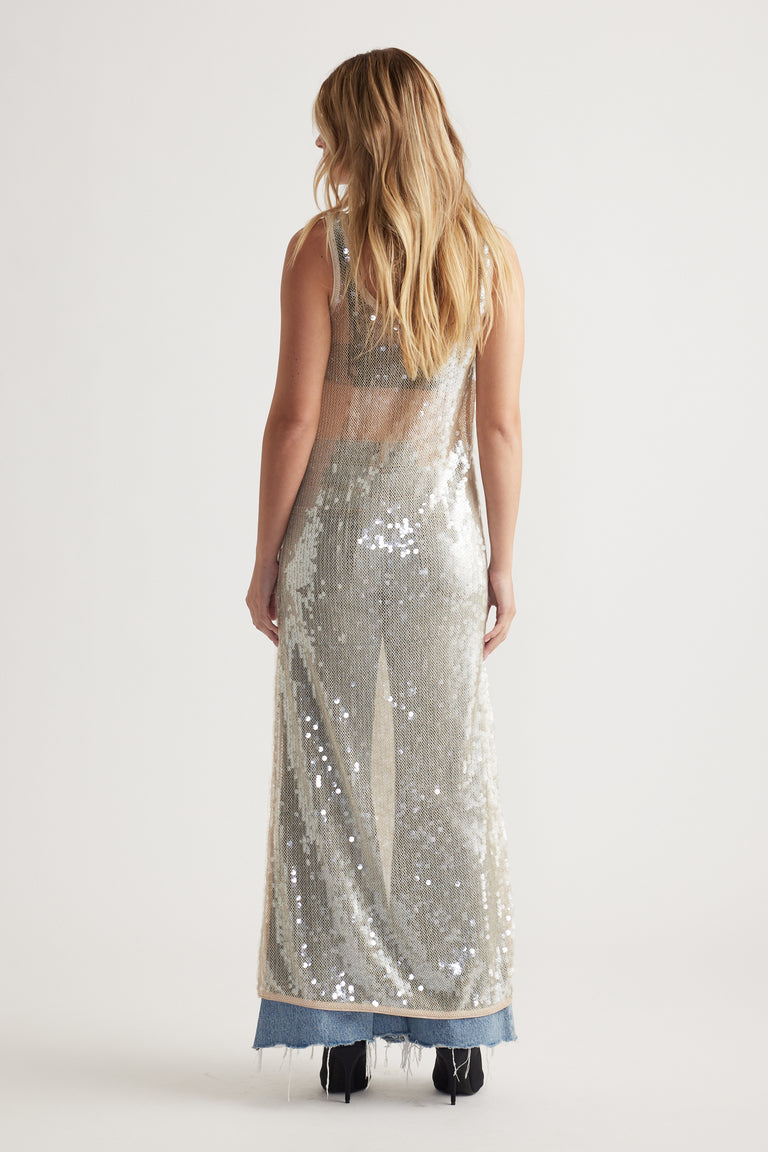 Woman wearing a sleeveless silver sequin maxi dress layered over raw-hem denim jeans and black ankle boots, back view