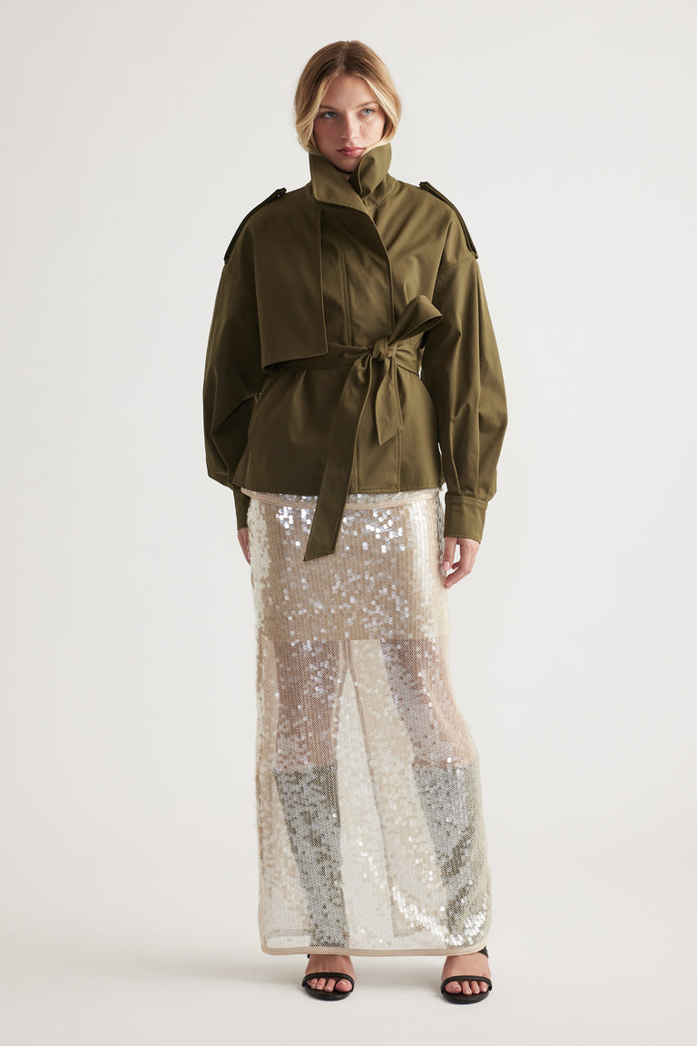 Woman wearing an olive belted jacket paired with a shimmering sequin maxi skirt and black open-toe heels