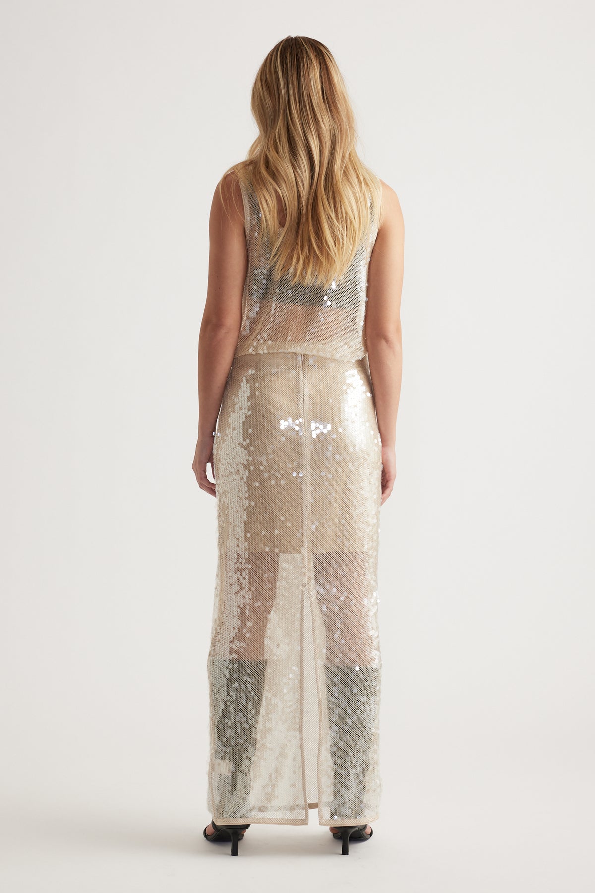 Back view of a woman wearing a shimmering champagne sequin maxi skirt with a sleeveless sequin top and black heels