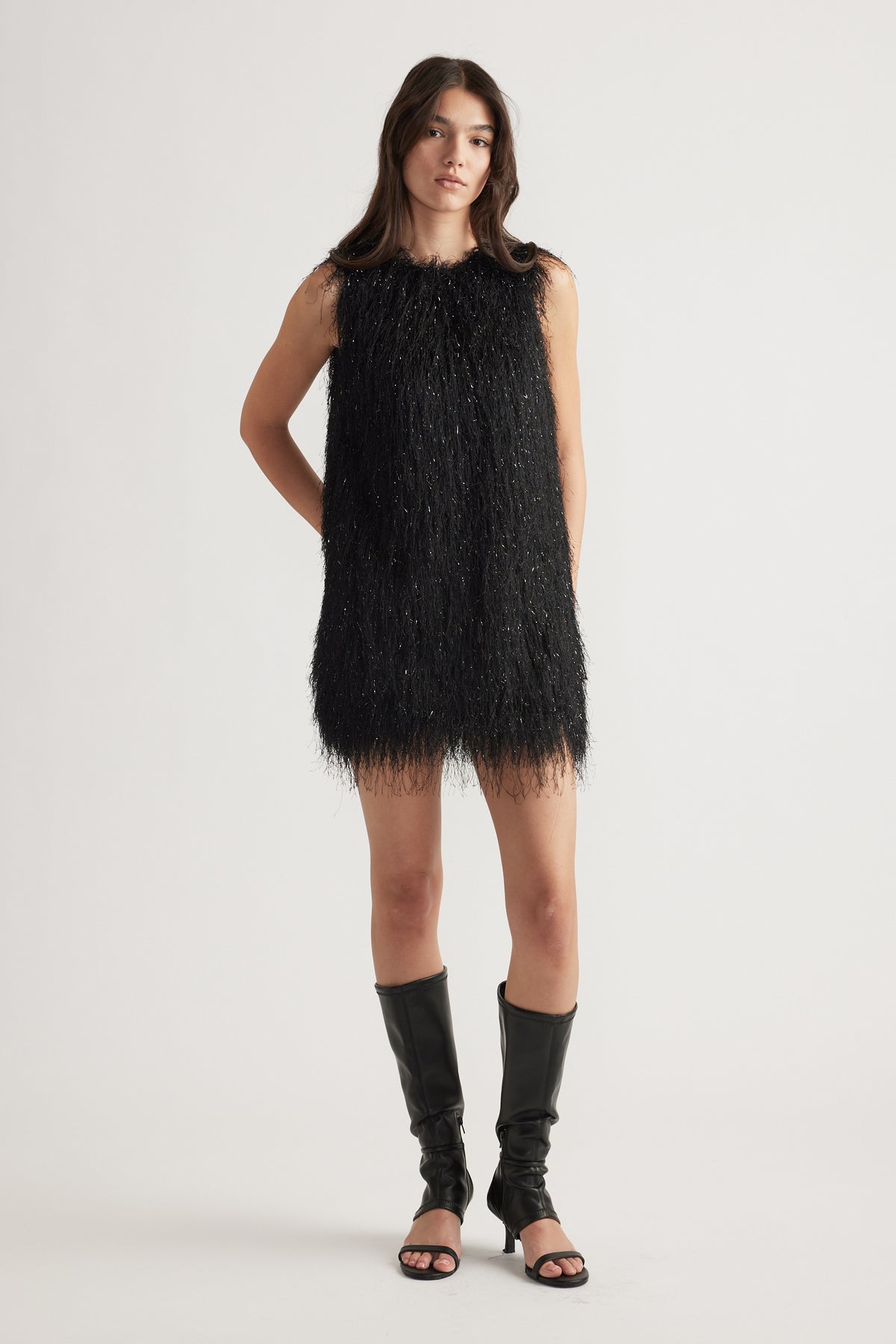 Woman wearing a black fringed mini dress paired with open-toe knee-high black leather boots, standing confidently