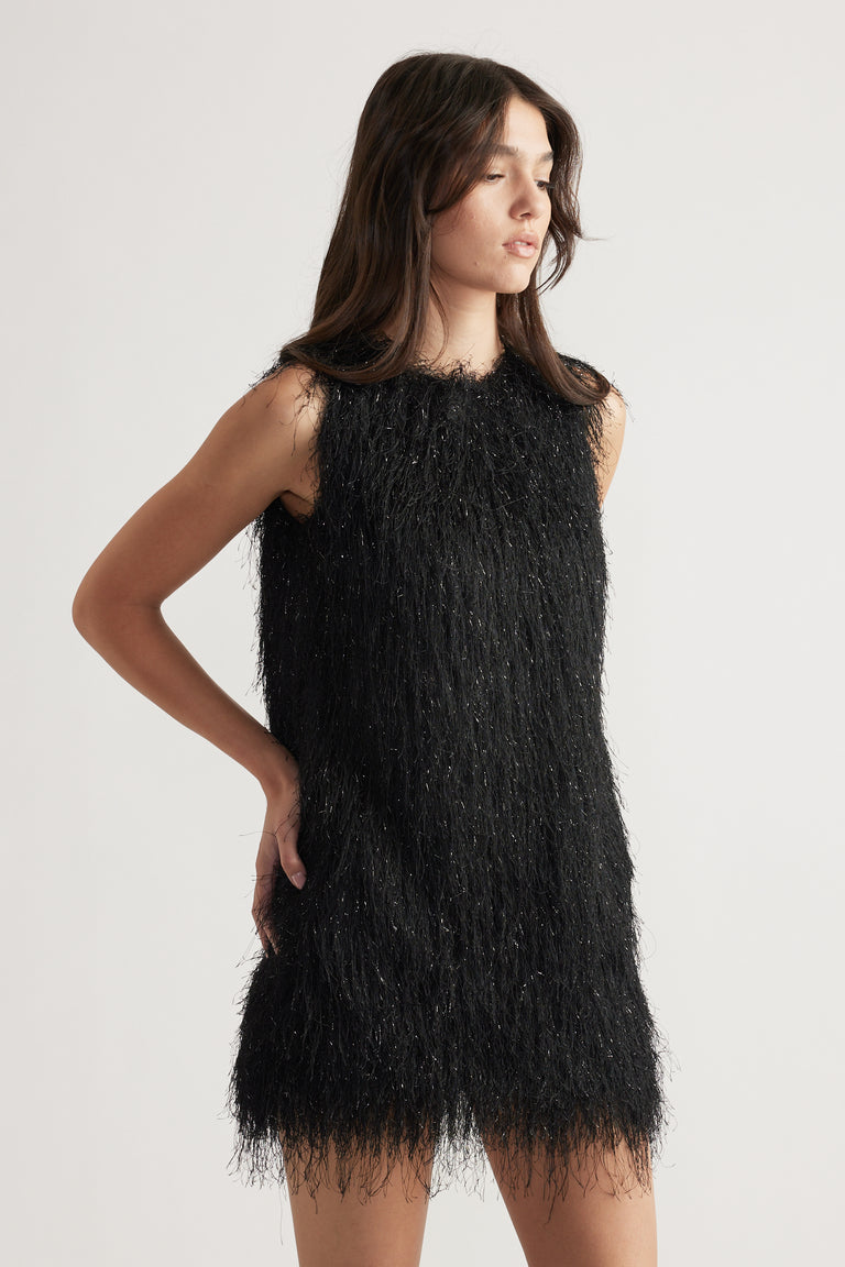 Woman wearing a sleeveless black mini dress with textured fringed details and subtle shimmer, posing with eyes closed.