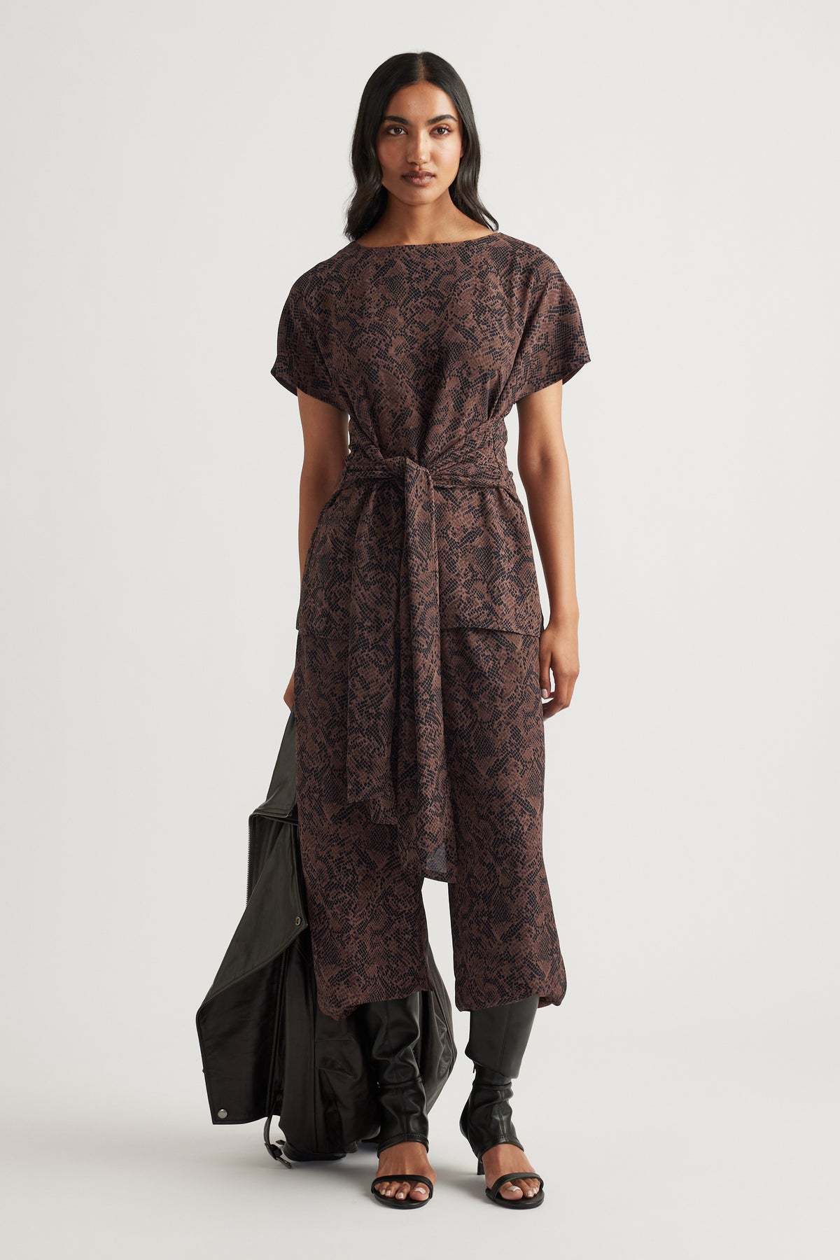 Woman wearing a brown snake-print tunic with a tie waist, styled with black pants and open-toe heels.