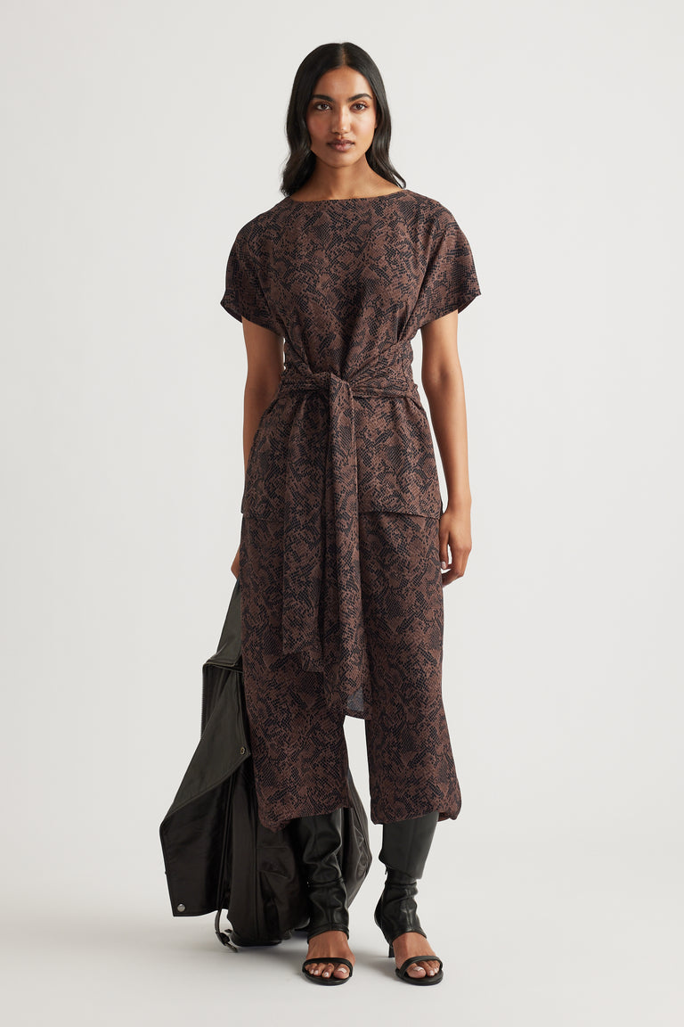 Woman wearing a brown snake-print tunic with a tie waist, styled with black pants and open-toe heels.