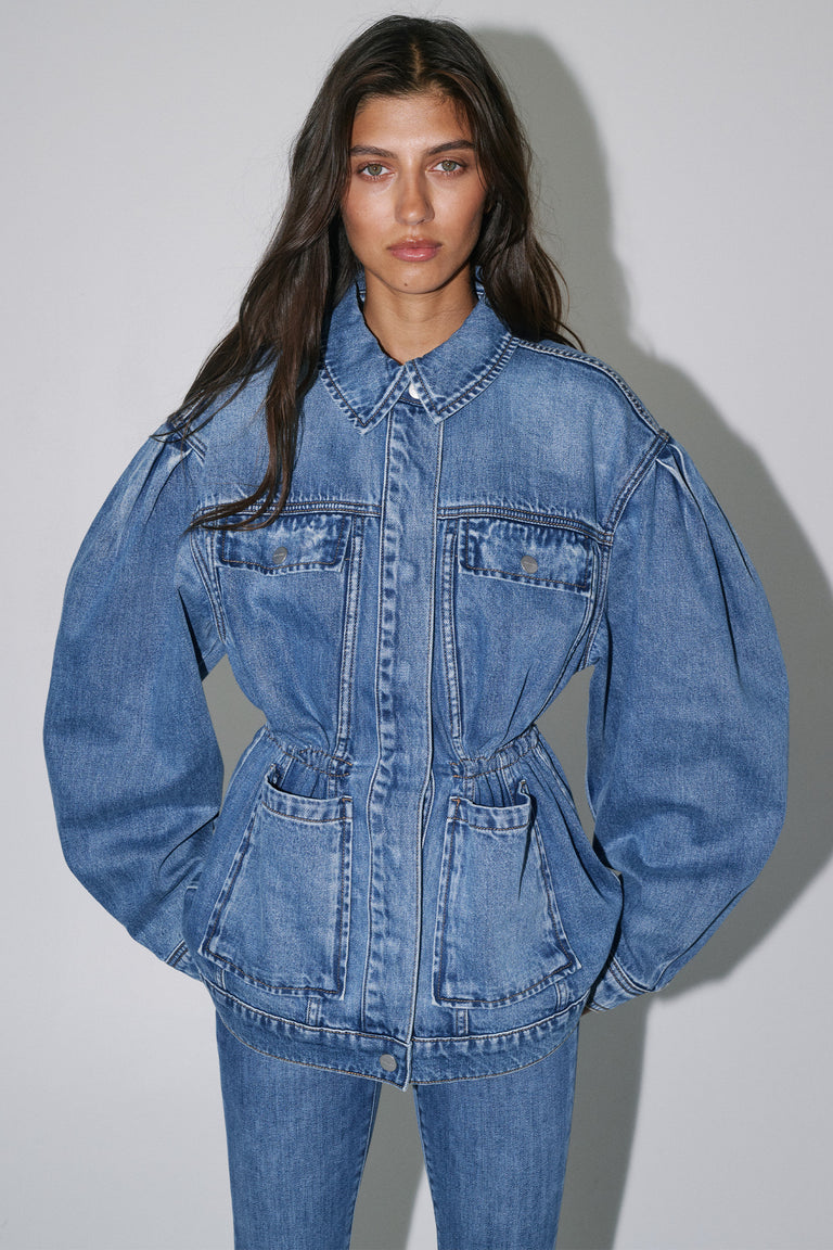 Woman wearing a cinched blue denim jacket with a collared neckline front pockets and matching jeans