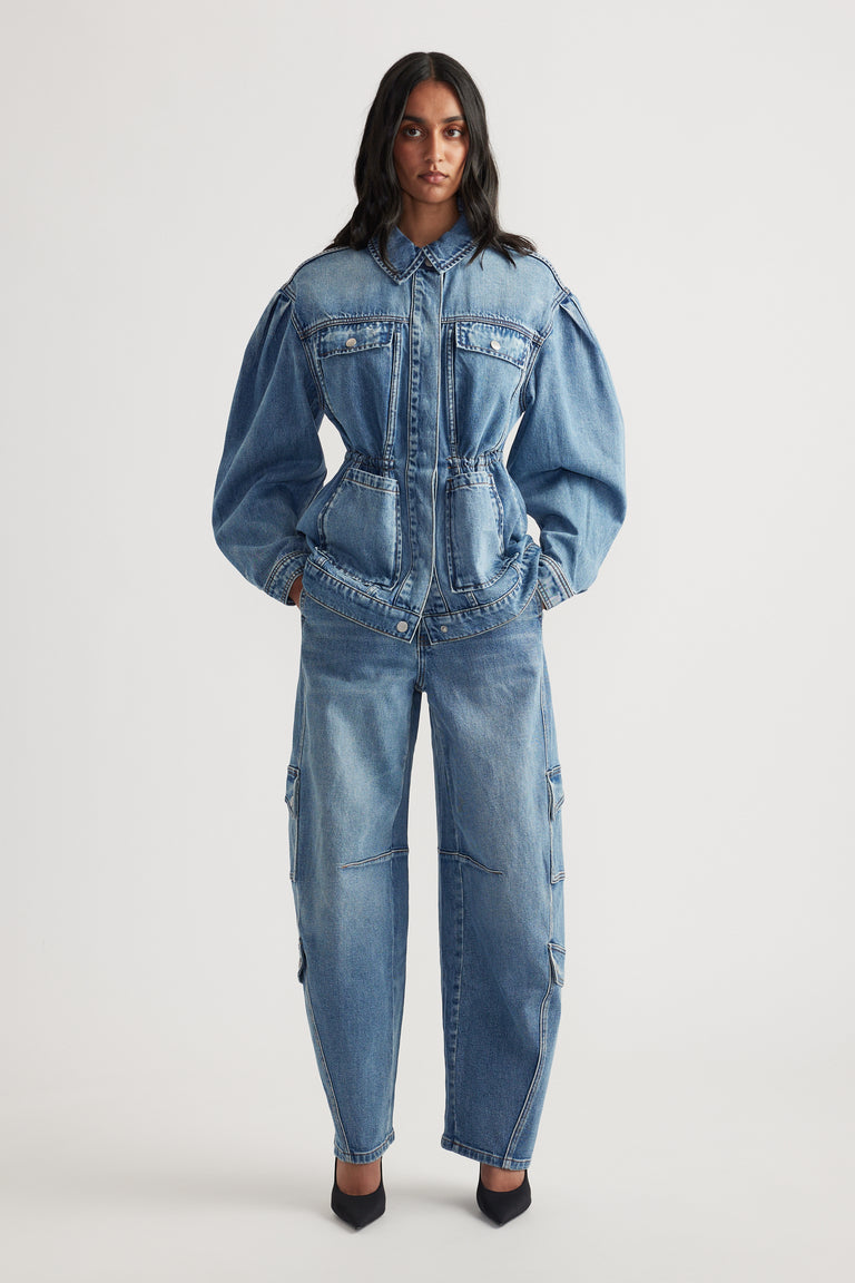 Front view of woman wearing long sleeve denim jacket with cinched waist paired with loose fit denim pants and black heels