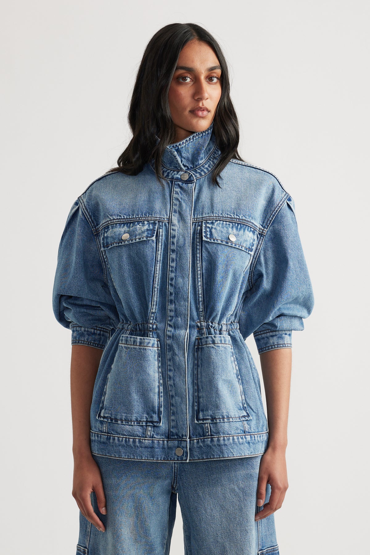 Front view of woman wearing a tailored denim jacket with puff sleeves and cinched waist paired with matching denim pants