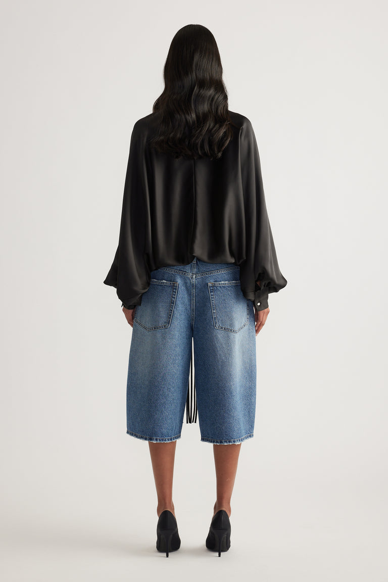 Back view of a model wearing a black satin top with batwing sleeves and a bubble hem, paired with blue denim shorts