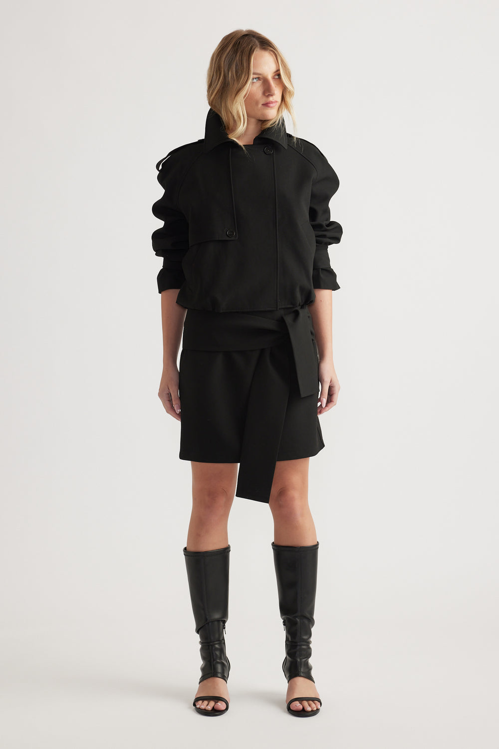 Woman wearing a black long-sleeve jacket and tie-detail mini skirt paired with tall open-toe boots on plain background