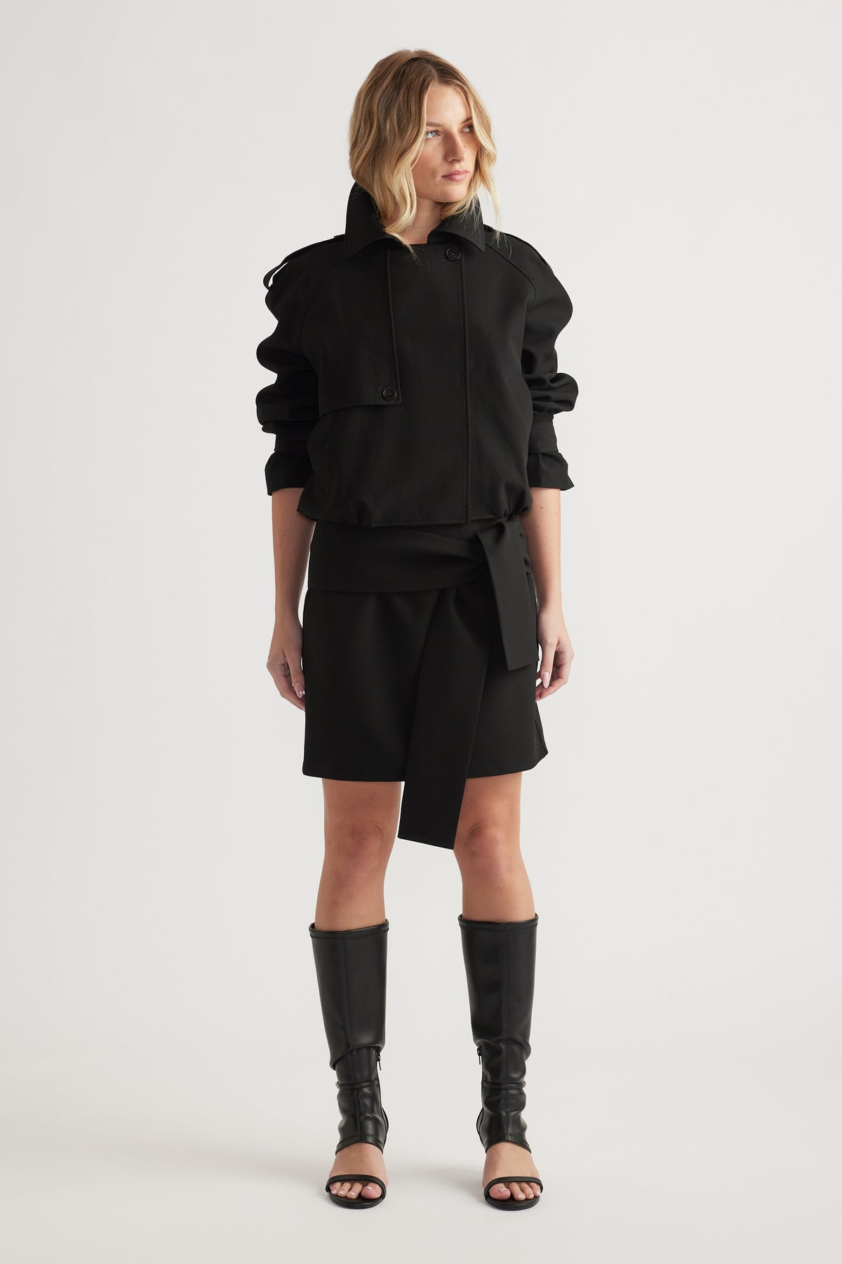 Woman wearing a black long-sleeve jacket and tie-detail mini skirt paired with tall open-toe boots on plain background