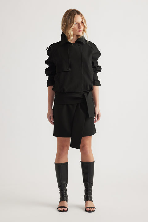 Woman wearing a black long-sleeve jacket and tie-detail mini skirt paired with tall open-toe boots on plain background