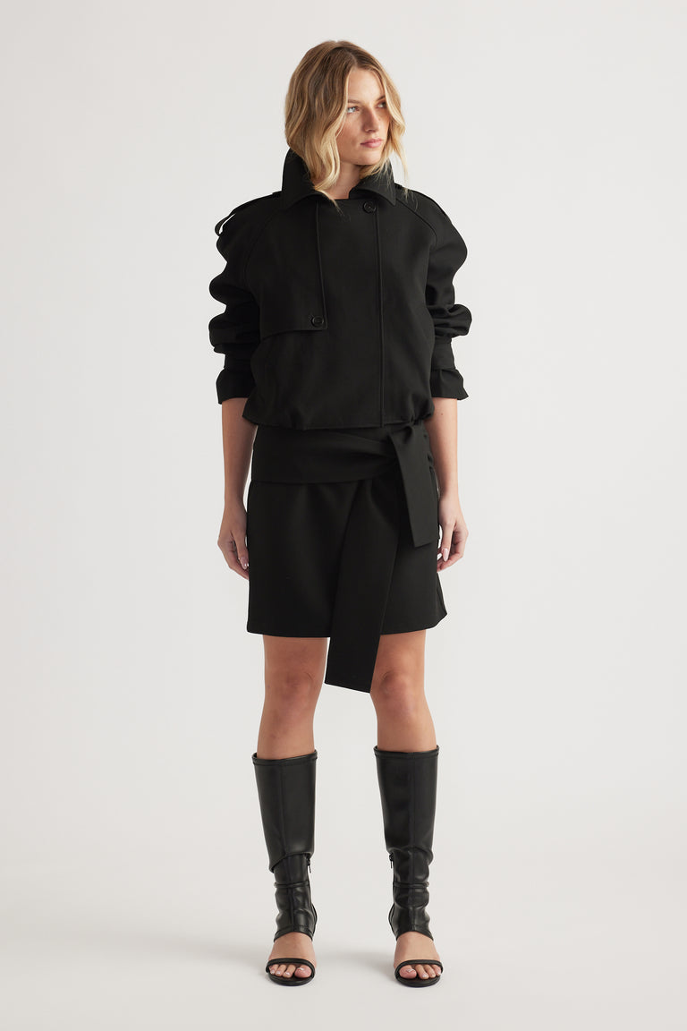 Woman wearing a black long-sleeve jacket and tie-detail mini skirt paired with tall open-toe boots on plain background
