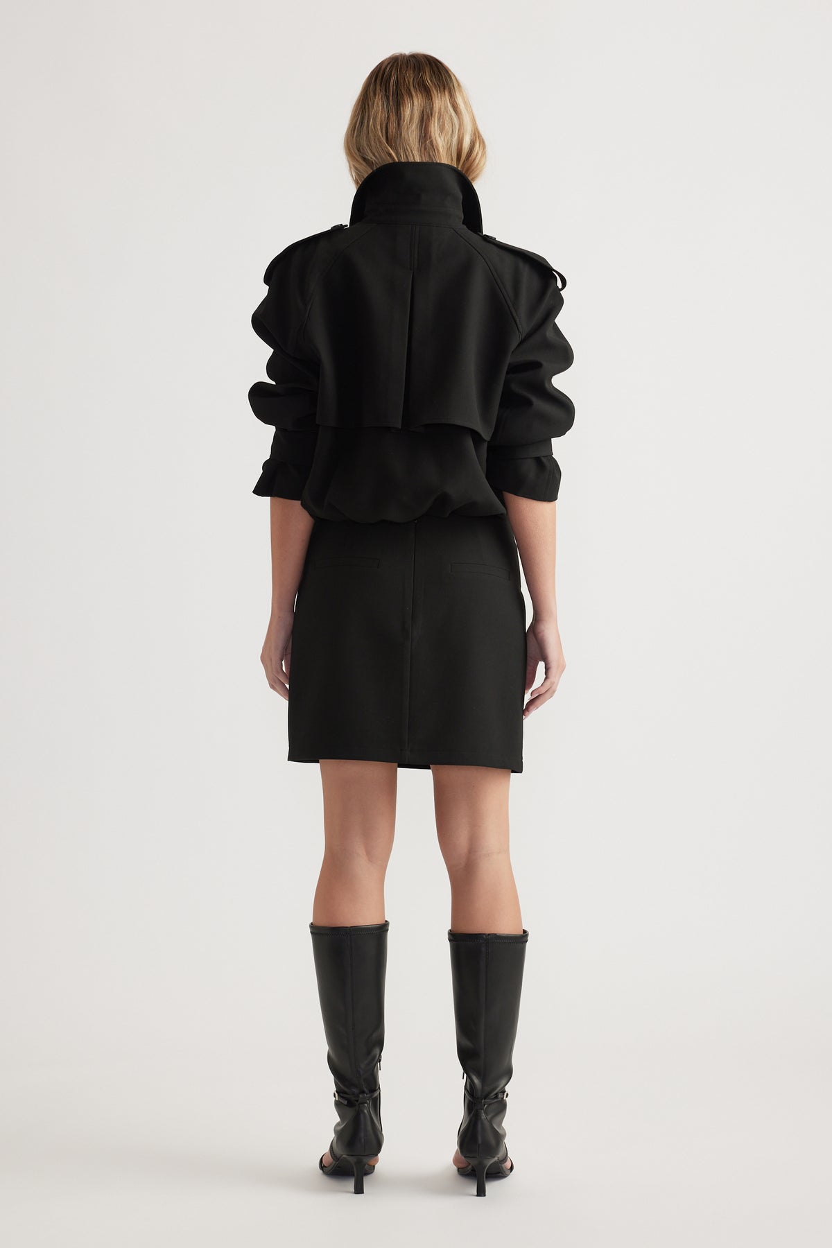 Woman wearing a black mini skirt and matching jacket with a high collar, styled with knee-high black boots, back view