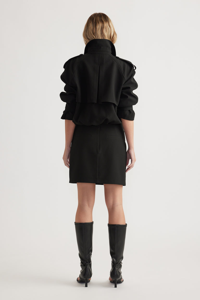 Woman wearing a black mini skirt and matching jacket with a high collar, styled with knee-high black boots, back view