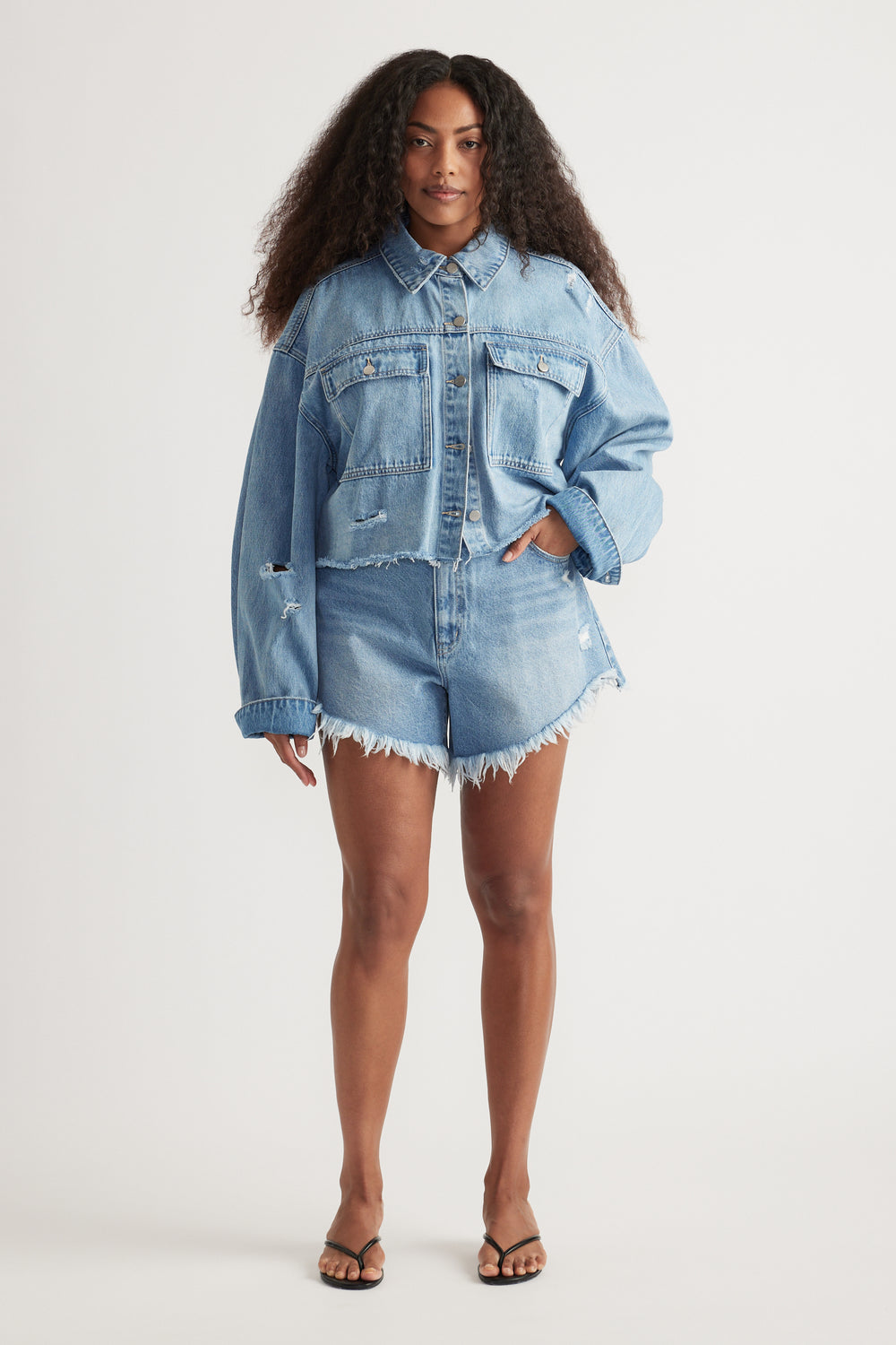 Woman wearing a cropped light blue distressed denim shirt with matching frayed shorts and sandals