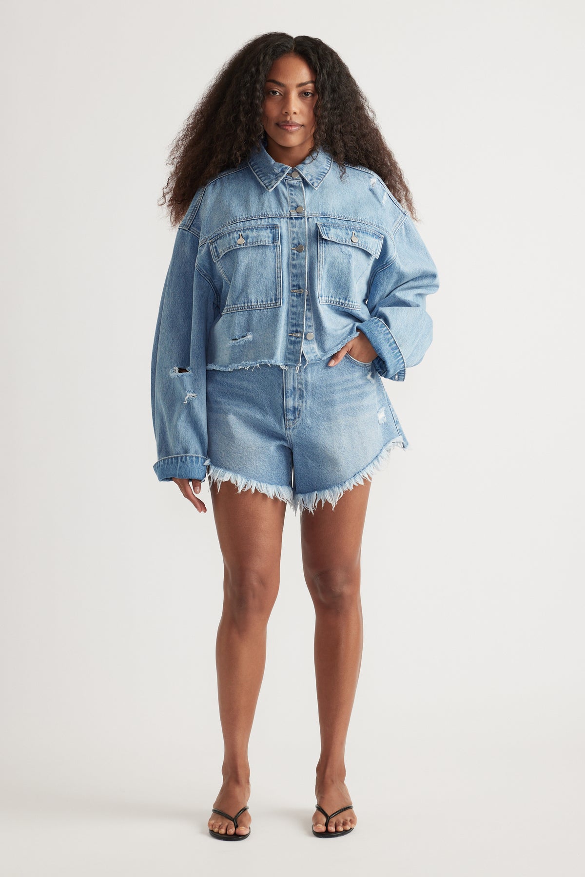 Woman wearing a cropped light blue distressed denim shirt with matching frayed shorts and sandals
