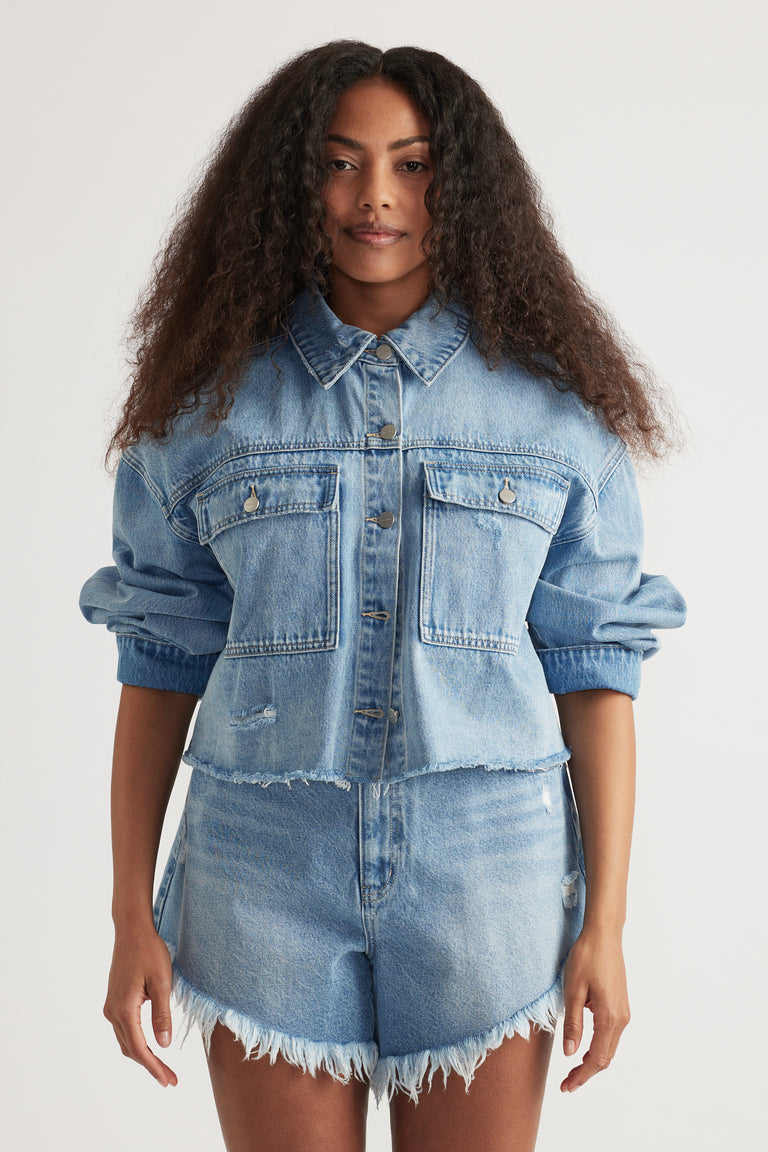 Woman wearing a cropped light wash denim shirt with rolled sleeves and matching frayed denim shorts