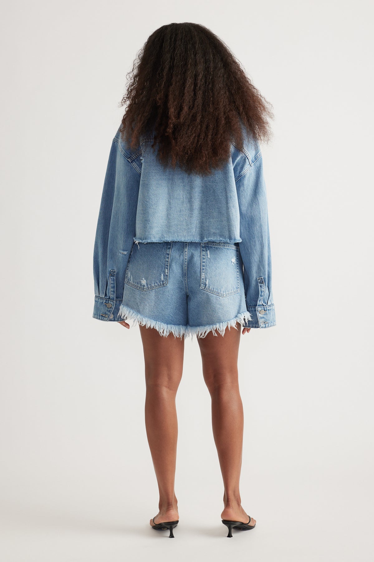 Back view of a woman wearing a cropped denim shirt with frayed denim shorts and black heeled sandals