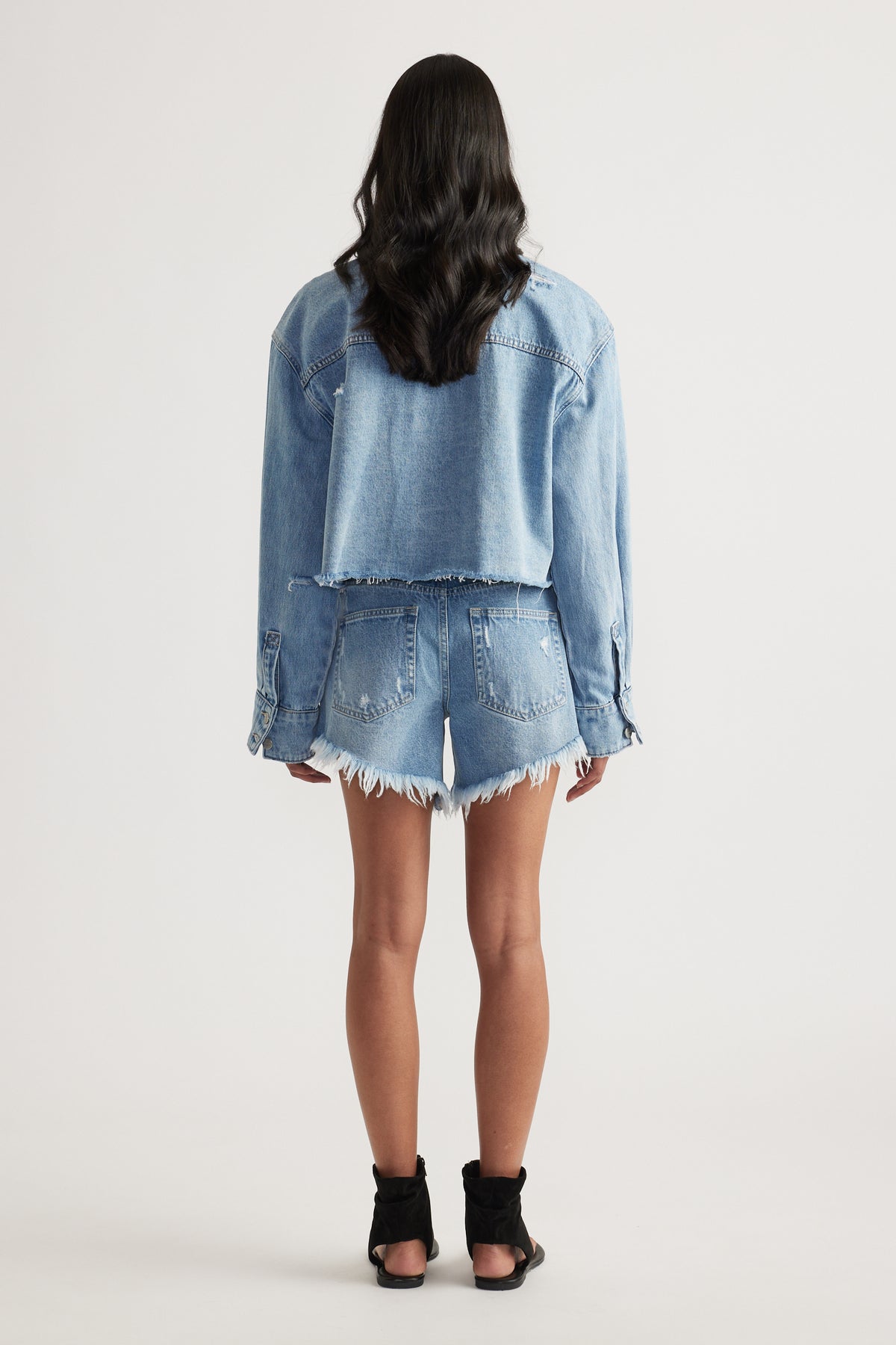 Back view of a woman wearing a cropped denim shirt, distressed denim shorts and black heeled sandals
