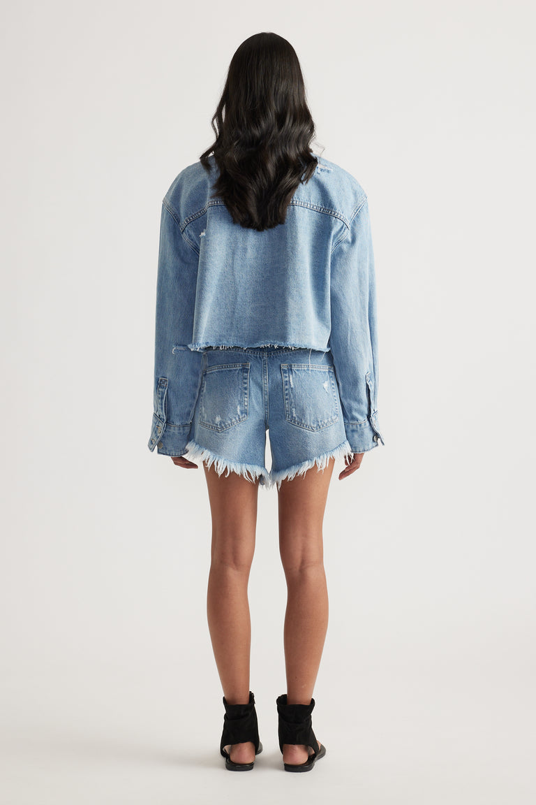 Back view of a woman wearing a cropped denim shirt, distressed denim shorts and black heeled sandals