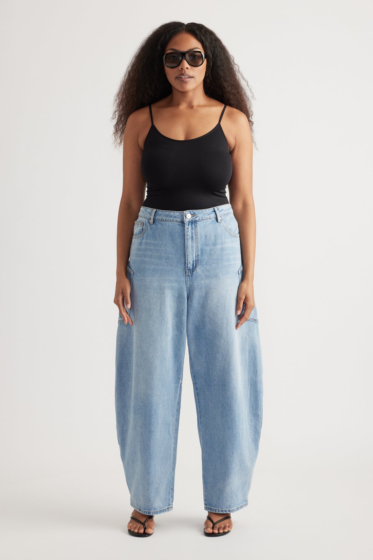 Woman wearing light wash barrel-leg cargo jeans with oversized pockets, a black camisole, sunglasses and sandals