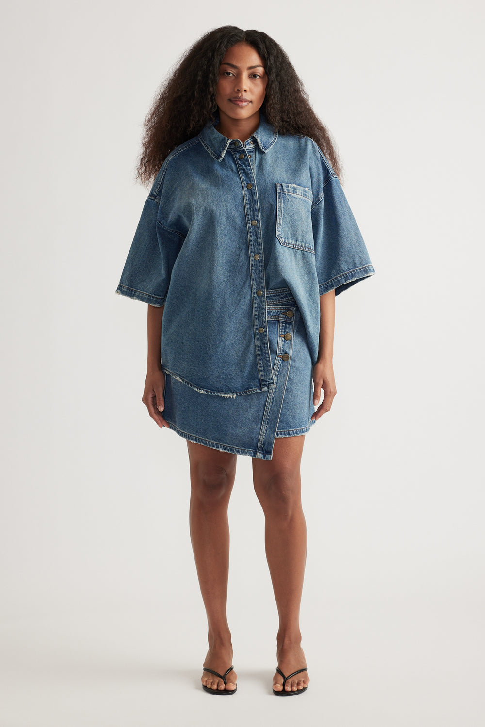 Woman wearing an oversized blue denim shirt with short sleeves, collar, front snaps and flat sandals