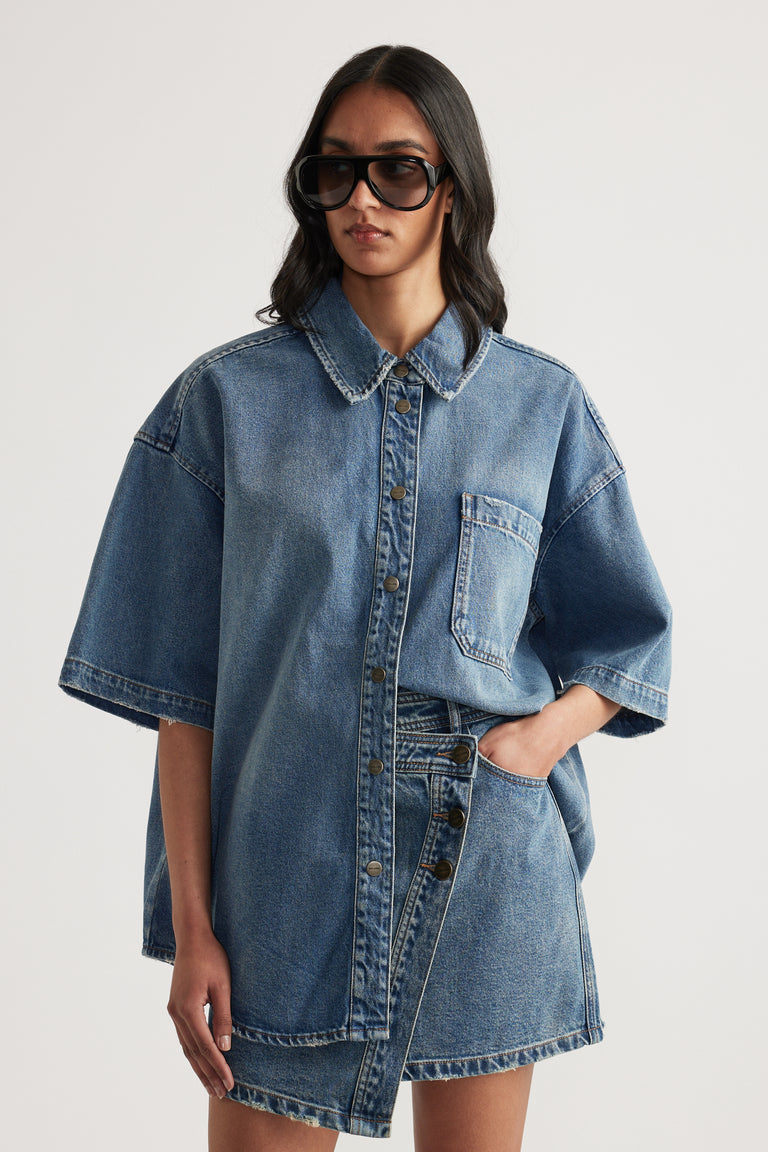 Woman wearing an oversized blue denim shirt with short sleeves, a collar, chest pocket and matching denim shorts