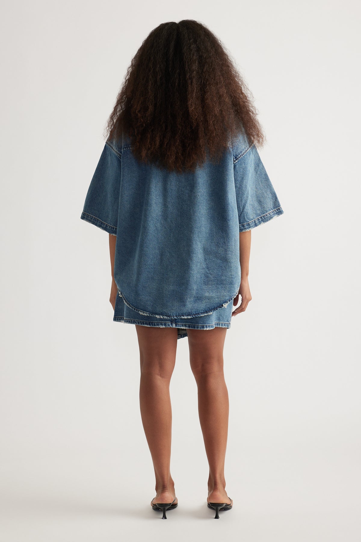Back view of a woman wearing an oversized blue denim shirt with short sleeves and heels
