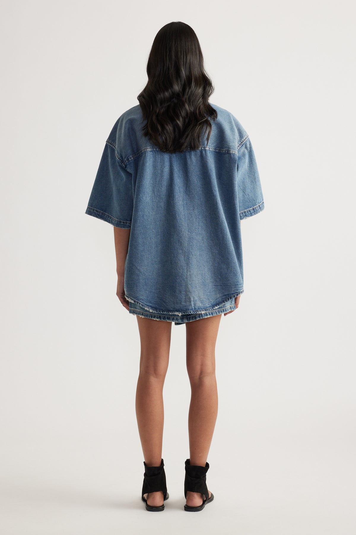 Back view of a model wearing an oversized blue denim shirt with short sleeves and black heeled sandals