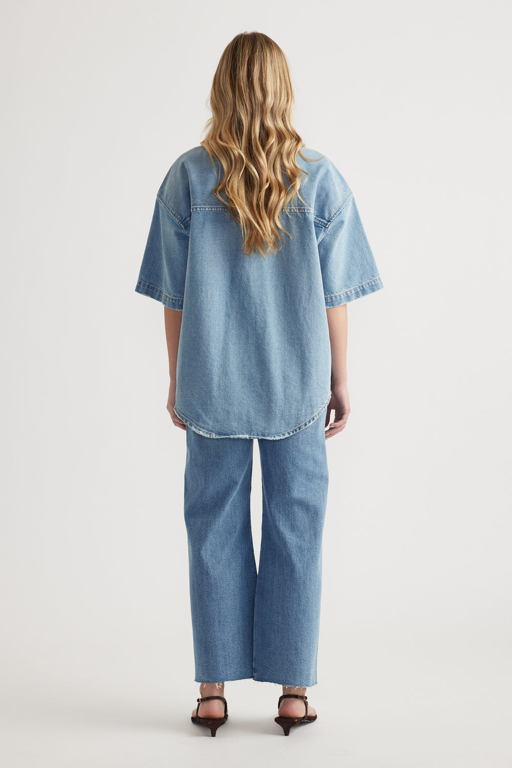 Back view of a woman wearing an oversized short sleeve denim shirt with wide leg jeans and heeled sandals